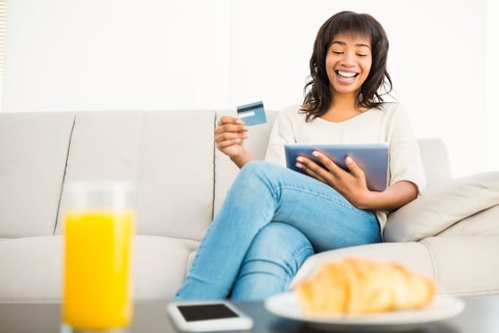 Woman on a couch, holding a tablet and a credt card in her hand.