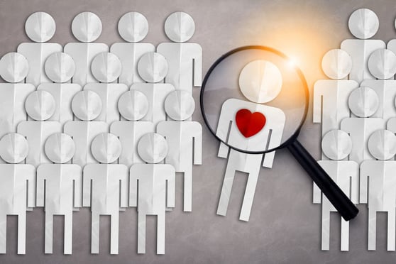 Graphic of a group of paper cut-out people, with a magnifying glass highlighting one person with a heart on their chest.