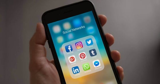 A hand holding a cell phone, showing 9 icons of different social media platforms.