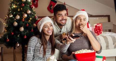 Image of three people in front of a Christmas tree, eating popcorn and holding a remote control.