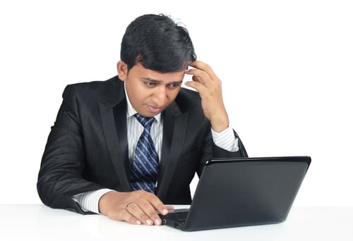 Image of man in suit, sitting in front of a laptop, scratching his head.