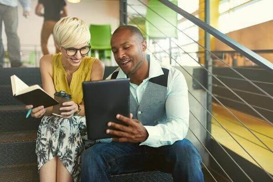 Two people sitting on stairs, looking at a tablet and smiling.