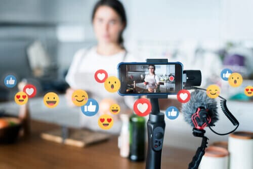 Woman livestreaming in the background, with phone on tripod in the foreground and flying emoji icons.