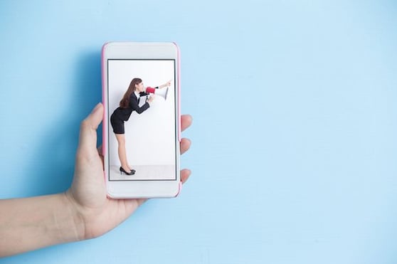 Hand holding a cellphone, with a woman speaking into a bullhorn.