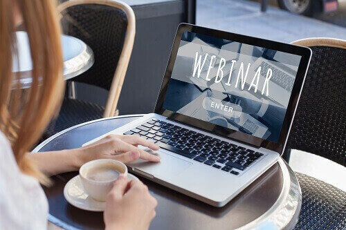 Woman with a cup of coffee sitting in front of a laptop, and the screen says 'Webinar Enter'.